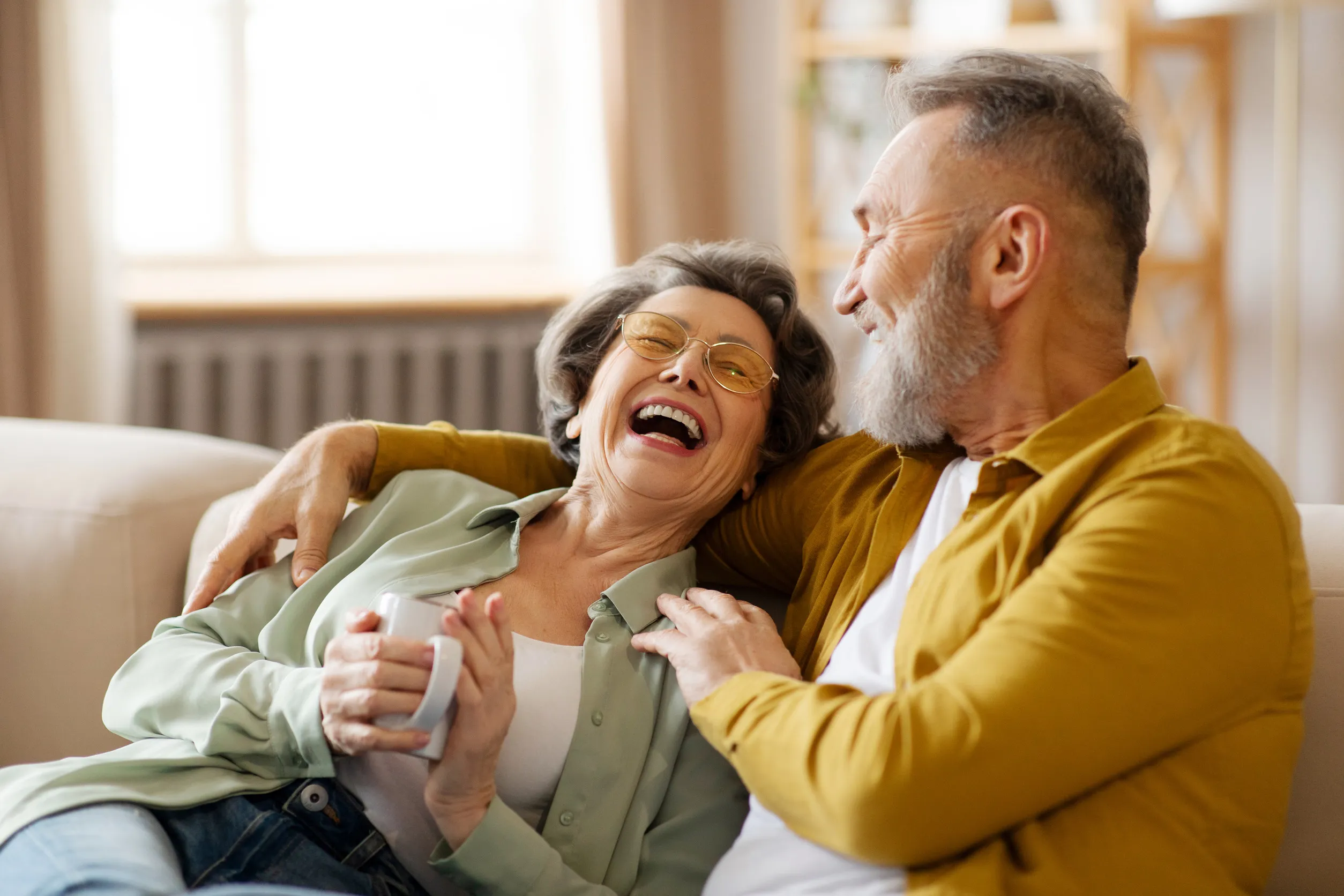 Senior spouses talking, sitting on sofa, woman holding cup of coffee and laughing to husband Senior spouses talking, sitting on sofa, woman holding cup of coffee and laughing to husband