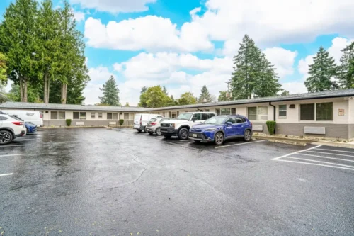 A parking lot with several vehicles, surrounded by single-story buildings and tall trees under a blue sky with fluffy clouds.