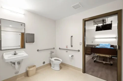 Accessible hospital bathroom with grab bars, a sink, toilet, and mirror. Adjacent room has a hospital bed. Clean and functional design.