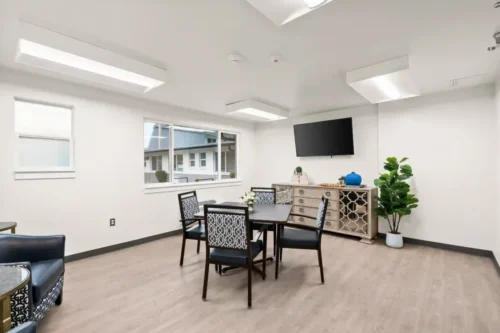 Bright, modern meeting room with wood flooring, decorative chairs around a table, a large TV, potted plant, and a sideboard beneath a wall-mounted TV.