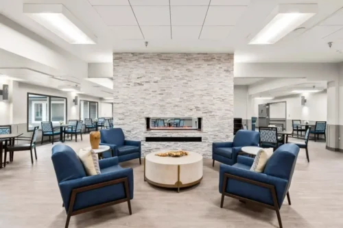 Modern lounge area with blue armchairs and a round table around a central stone fireplace. Light and airy, with a calm, inviting atmosphere.