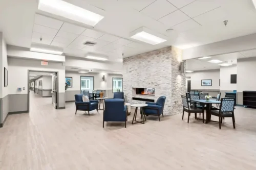 Spacious, brightly lit lounge featuring blue armchairs and round tables. A stone fireplace is central, creating a cozy and inviting atmosphere.